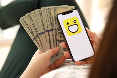 KYIV, UKRAINE - 1 Nisan 2024 Hago simgesi akıllı telefon ekranında ve kadın elinde para. Uygulama logolu iPhone görüntüsü ve kızların elinde 100 dolarlık banknotlar