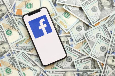 KYIV, UKRAINE - 1 Nisan 2024 Facebook simgesi, akıllı telefon ekranı ve birçok kullanım parası. Uygulama logolu iPhone görüntüsü ve bir sürü yüz dolarlık banknotlar
