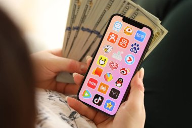 KYIV, UKRAINE - Nisan 1, 2024 Asya uygulama simgesi akıllı telefon ekranında ve kadın elinde para. Uygulama logolu iPhone görüntüsü ve kızların elinde 100 dolarlık banknotlar