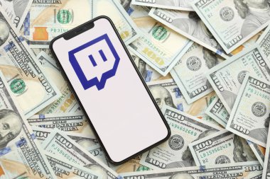 KYIV, UKRAINE - 1 Nisan 2024 Twitch simgesi, akıllı telefon ekranı ve birçok kullanım parası. Uygulama logolu iPhone görüntüsü ve bir sürü yüz dolarlık banknotlar