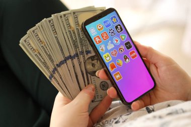 KYIV, UKRAINE - Nisan 1, 2024 Asya uygulama simgesi akıllı telefon ekranında ve kadın elinde para. Uygulama logolu iPhone görüntüsü ve kızların elinde 100 dolarlık banknotlar