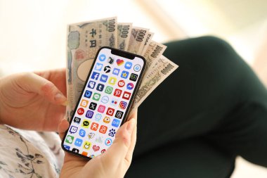 KYIV, UKRAINE - Nisan 1, 2024 Akıllı telefon ekranında birçok uygulama simgesi ve kadın elinde para. Uygulama logosu ve Japon Yen 'i olan iPhone ekranı kadınların elinde. Kapatın.