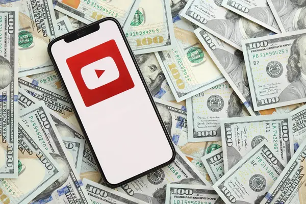 KYIV, UKRAINE - Nisan 1, 2024 Youtube simgesi, akıllı telefon ekranı ve birçok kullanım parası. Uygulama logolu iPhone görüntüsü ve bir sürü yüz dolarlık banknotlar