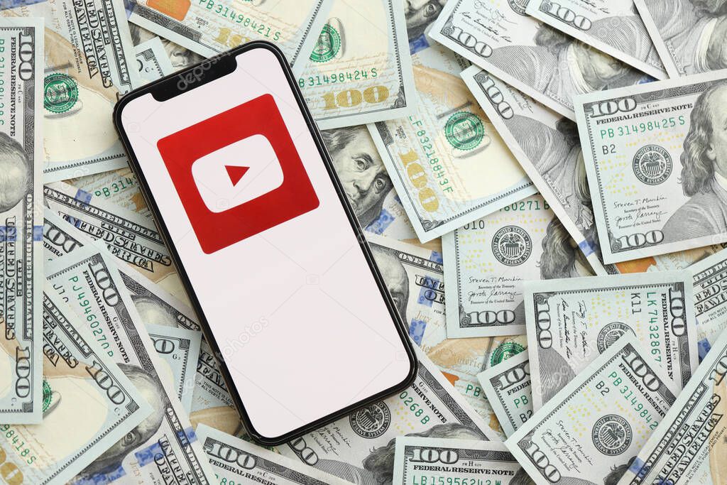 KYIV, UKRAINE - APRIL 1, 2024 Youtube icon on smartphone screen and many usd money bills. iPhone display with app logo and lot of hundred dollars banknotes close up