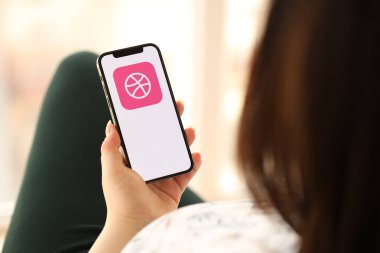 KYIV, UKRAINE - Nisan 1, 2024 Kapalı alandaki genç kadınların elinde akıllı telefon ekranında Dribbble simgesi. iPhone uygulaması kullanan kadın