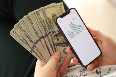 KYIV, UKRAINE - 1 Nisan 2024 Oasis simgesi akıllı telefon ekranında ve kadın elinde para. Uygulama logolu iPhone görüntüsü ve kızların elinde 100 dolarlık banknotlar