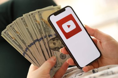 KYIV, UKRAINE - Nisan 1, 2024 Youtube simgesi akıllı telefon ekranında ve kadın elinde para. Uygulama logolu iPhone görüntüsü ve kızların elinde 100 dolarlık banknotlar