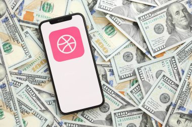 KYIV, UKRAINE - Nisan 1, 2024 Akıllı telefon ekranında Dribbble simgesi ve birçok kullanım dışı para kaynağı. Uygulama logolu iPhone görüntüsü ve bir sürü yüz dolarlık banknotlar