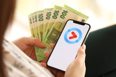 KYIV, UKRAINE - 1 Nisan 2024 Youku simgesi akıllı telefon ekranında ve kadın elinde para. Uygulama logosuna sahip iPhone görüntüsü ve kızların elinde 100 Euro 'luk banknot