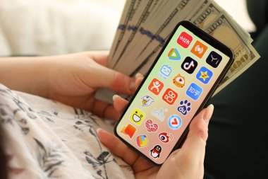 KYIV, UKRAINE - Nisan 1, 2024 Asya uygulama simgesi akıllı telefon ekranında ve kadın elinde para. Uygulama logolu iPhone görüntüsü ve kızların elinde 100 dolarlık banknotlar