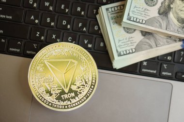 KYIV, UKRAINE - AUGUST 17, 2024 TRON Coin with big amount of hundred US dollar bills on laptop surface close up clipart