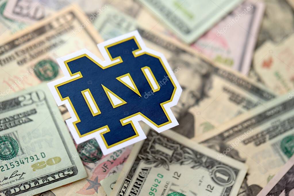 KYIV, UKRAINE - FEBRUARY 23, 2025 Notre Dame Fighting Irish basketball team badge lies on big amount of US dollar bills close up