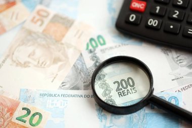 Brezilya reais banknotları ve büyüteçli hesap makinesi. Mevcut Brezilya gerçek para faturaları ve loupe ile calc büyük miktarda masada yatıyor. Brezilya 'da vergilendirme ve para konsepti
