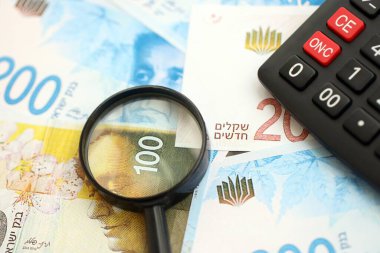Israel New Shekels banknotları ve büyüteçle birlikte hesap makinesi. Mevcut İsrail para faturaları ve loupe ile calc büyük miktarda masada yatıyor. İsrail 'de vergilendirme ve para kavramı