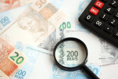 Brezilya reais banknotları ve büyüteçli hesap makinesi. Mevcut Brezilya gerçek para faturaları ve loupe ile calc büyük miktarda masada yatıyor. Brezilya 'da vergilendirme ve para konsepti