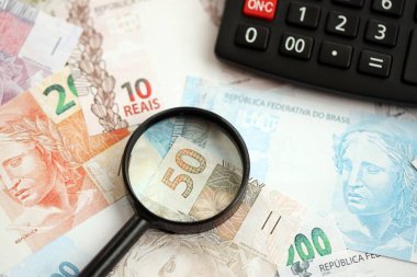 Brezilya reais banknotları ve büyüteçli hesap makinesi. Mevcut Brezilya gerçek para faturaları ve loupe ile calc büyük miktarda masada yatıyor. Brezilya 'da vergilendirme ve para konsepti