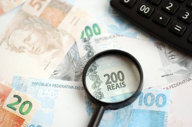 Brezilya reais banknotları ve büyüteçli hesap makinesi. Mevcut Brezilya gerçek para faturaları ve loupe ile calc büyük miktarda masada yatıyor. Brezilya 'da vergilendirme ve para konsepti