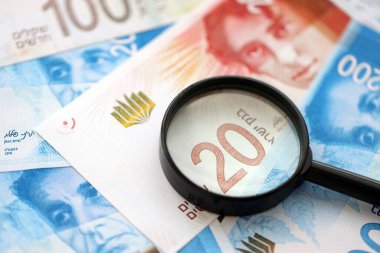 Israel New Shekels banknotları ve büyüteç. Şu anki İsrail 'in yüksek meblağlı faturaları masanın üzerinde yatıyor. İsrail 'de mali izleme ve vergilendirme kavramı