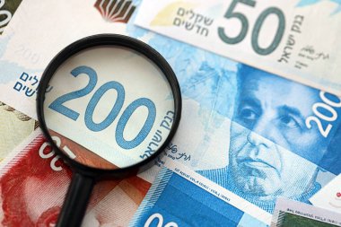 Israel New Shekels banknotları ve büyüteç. Şu anki İsrail 'in yüksek meblağlı faturaları masanın üzerinde yatıyor. İsrail 'de mali izleme ve vergilendirme kavramı