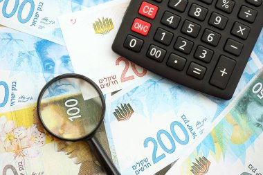 Israel New Shekels banknotları ve büyüteçle birlikte hesap makinesi. Mevcut İsrail para faturaları ve loupe ile calc büyük miktarda masada yatıyor. İsrail 'de vergilendirme ve para kavramı