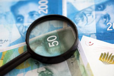 Israel New Shekels banknotları ve büyüteç. Şu anki İsrail 'in yüksek meblağlı faturaları masanın üzerinde yatıyor. İsrail 'de mali izleme ve vergilendirme kavramı