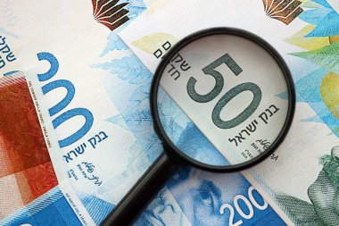 Israel New Shekels banknotları ve büyüteç. Şu anki İsrail 'in yüksek meblağlı faturaları masanın üzerinde yatıyor. İsrail 'de mali izleme ve vergilendirme kavramı