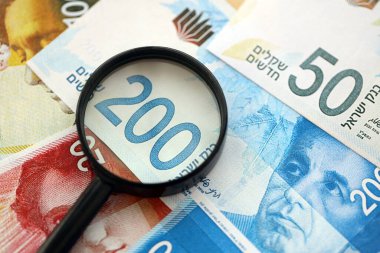 Israel New Shekels banknotları ve büyüteç. Şu anki İsrail 'in yüksek meblağlı faturaları masanın üzerinde yatıyor. İsrail 'de mali izleme ve vergilendirme kavramı
