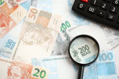 Brezilya reais banknotları ve büyüteçli hesap makinesi. Mevcut Brezilya gerçek para faturaları ve loupe ile calc büyük miktarda masada yatıyor. Brezilya 'da vergilendirme ve para konsepti