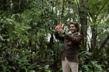 Şehirdeki yeşil bir parkta gündüz vakti akıllı telefonlu genç bir adamın görüntüsü. Akıllı telefonla selfie çekiyorum..
