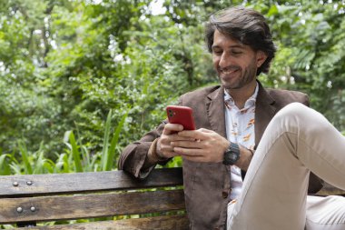 Akıllı telefonlu bir gencin gündüz vakti şehirdeki yeşil bir parkta bankta oturduğunu görmek. Yüksek kalite fotoğraf. 