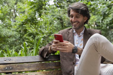 Akıllı telefonlu bir gencin gündüz vakti şehirdeki yeşil bir parkta bankta oturduğunu görmek. Yüksek kalite fotoğraf. 