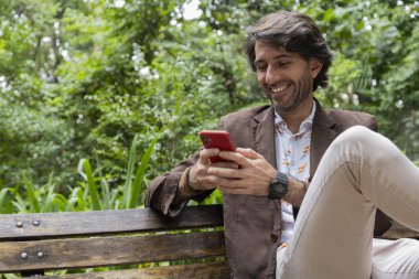 Akıllı telefonlu bir gencin gündüz vakti şehirdeki yeşil bir parkta bankta oturduğunu görmek. Yüksek kalite fotoğraf. 