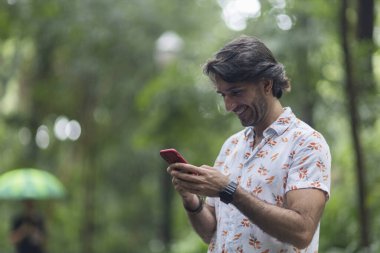 Şehirdeki yeşil bir parkta gündüz vakti akıllı telefonlu genç bir adam görülüyor..