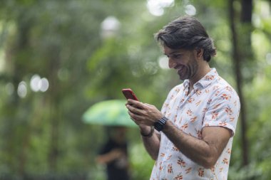 Şehirdeki yeşil bir parkta gündüz vakti akıllı telefonlu genç bir adam görülüyor..