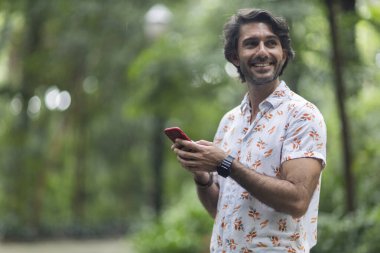 Şehirdeki yeşil bir parkta gündüz vakti akıllı telefonlu genç bir adam görülüyor..