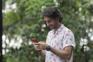 Şehirdeki yeşil bir parkta gündüz vakti akıllı telefonlu genç bir adam görülüyor..