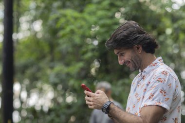 Şehirdeki yeşil bir parkta gündüz vakti akıllı telefonlu genç bir adam görülüyor..