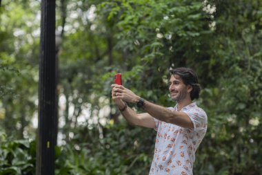 Şehirdeki yeşil bir parkta gündüz vakti akıllı telefonlu genç bir adamın görüntüsü. Akıllı telefonla selfie çekiyorum..