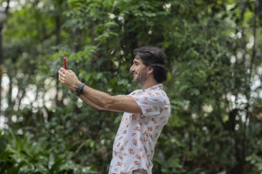 Şehirdeki yeşil bir parkta gündüz vakti akıllı telefonlu genç bir adamın görüntüsü. Akıllı telefonla selfie çekiyorum..