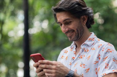 Şehirdeki yeşil bir parkta gündüz vakti akıllı telefonlu genç bir adam görülüyor..