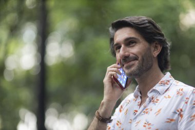 Şehirdeki yeşil bir parkta gündüz vakti akıllı telefonlu genç bir adam görülüyor..