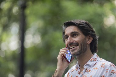 Şehirdeki yeşil bir parkta gündüz vakti akıllı telefonlu genç bir adam görülüyor..