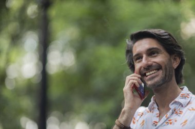 Şehirdeki yeşil bir parkta gündüz vakti akıllı telefonlu genç bir adam görülüyor..