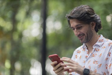 Şehirdeki yeşil bir parkta gündüz vakti akıllı telefonlu genç bir adam görülüyor..