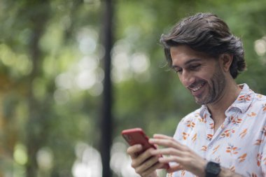 Şehirdeki yeşil bir parkta gündüz vakti akıllı telefonlu genç bir adam görülüyor..