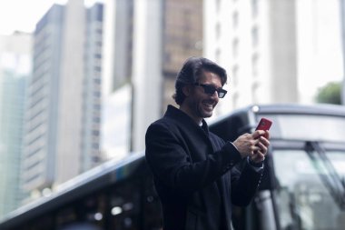 Gündüz vakti güneş gözlüklü ve akıllı telefonlu arka planda şehir manzaralı iyi giyimli genç bir adamın görüntüsü. Yüksek kalite fotoğraf. 