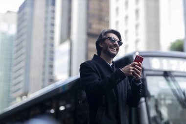 Gündüz vakti güneş gözlüklü ve akıllı telefonlu arka planda şehir manzaralı iyi giyimli genç bir adamın görüntüsü. Yüksek kalite fotoğraf. 