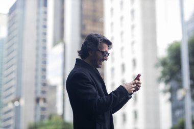 Gündüz vakti güneş gözlüklü ve akıllı telefonlu arka planda şehir manzaralı iyi giyimli genç bir adamın görüntüsü. Yüksek kalite fotoğraf. 