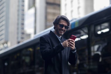 Gündüz vakti güneş gözlüklü ve akıllı telefonlu arka planda şehir manzaralı iyi giyimli genç bir adamın görüntüsü. Yüksek kalite fotoğraf. 