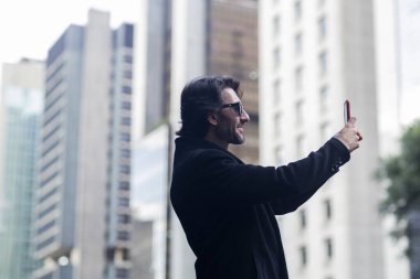 Gündüz vakti güneş gözlüklü ve akıllı telefonlu arka planda şehir manzaralı iyi giyimli genç bir adamın görüntüsü. Akıllı telefonla selfie çekiyorum..